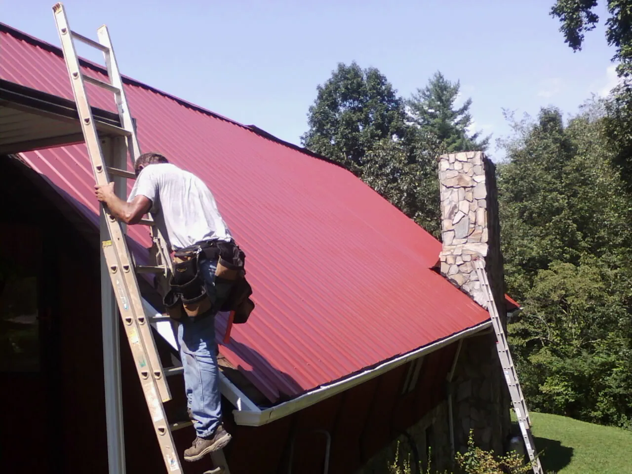 Roofer on a ladder beside a red metal roof and stone chimney — Soffit & Fascia Repair by Roofing Brothers in Beresford
